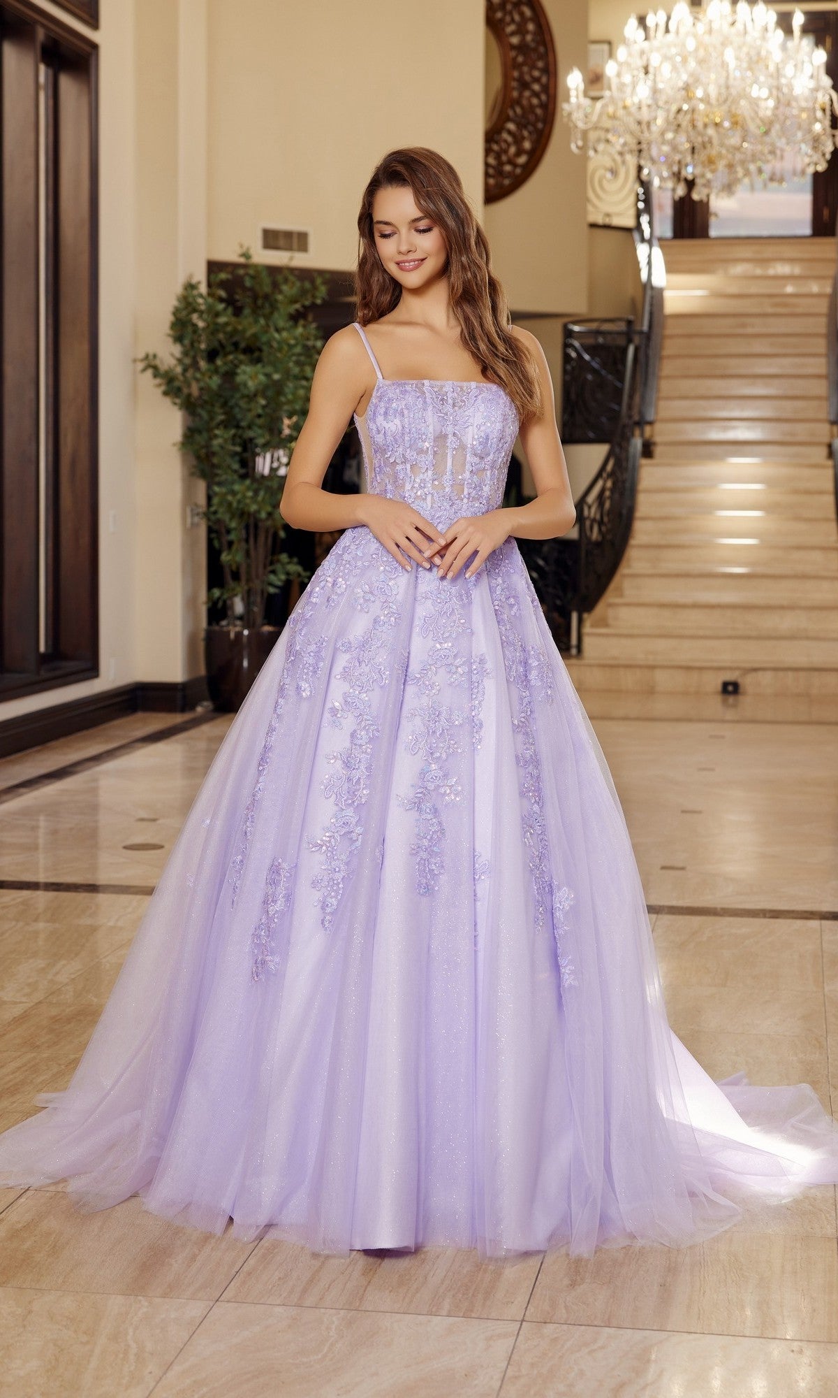 A woman stands indoors by a staircase and chandelier, wearing the Nox Anabel H1880 Long Prom Dress in lilac, featuring a floor-length a-line skirt, floral embroidery, and a fitted bodice.